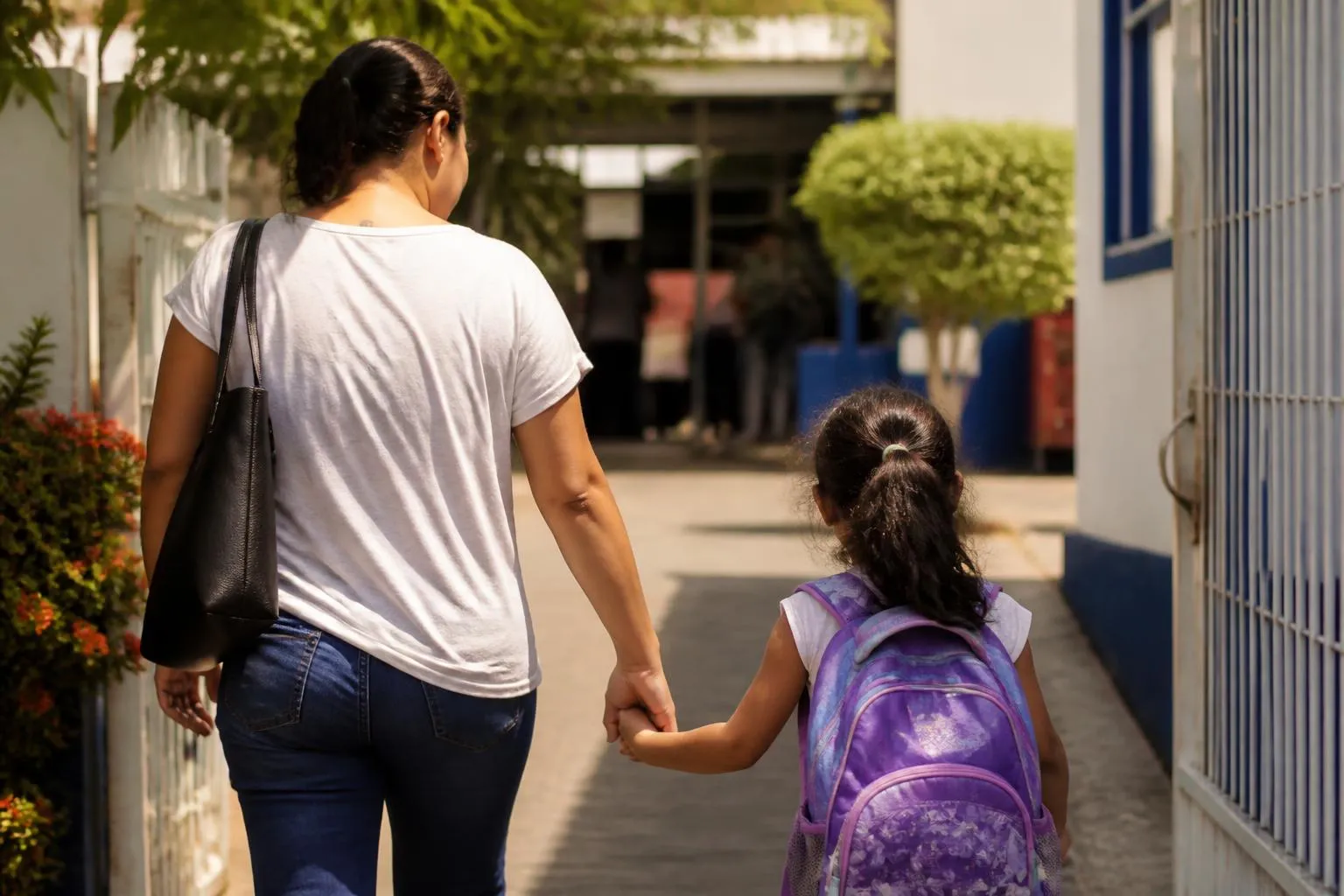Mãe levando a filha pela mão até a escola, mochila roxa nas costas